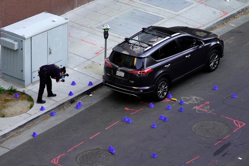 A police office photographs evidence on a road near an SUV. Many small blue cones are on the road and footpath.