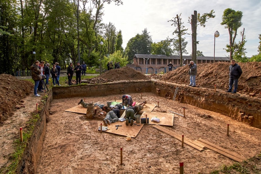 Inside a large excavation pit, five archaeologists can be seen on hands and knees digging something out.