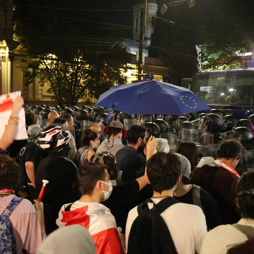 A crowd of people in civilian clothes and Georgian flags facing a crowd of heavy duty police