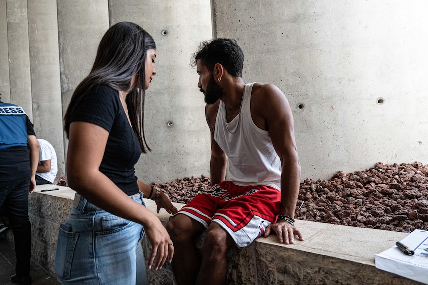 A young woman speaks to a young man sitting on a wall, whose face is turned away from the camera
