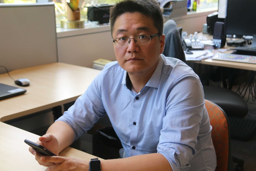 A man sits at a desk holding a mobile phone.