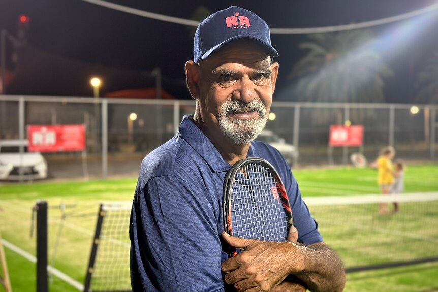 A man wearing a baseball cap holds a tennis racquet to his chest under the lights of a tennis court at night 