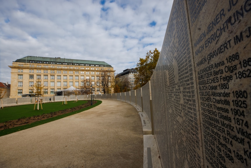 A wall is seen with names on it, while behind it a park and large building are visible. 