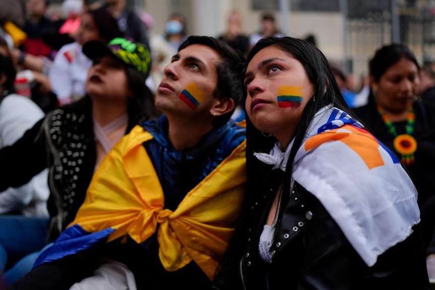 A man in a woman sit in a crowd with the Colombia flag painted on their faces.