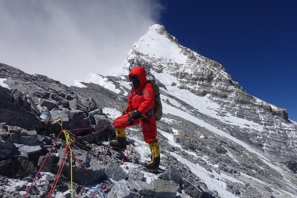 Man in red suit standing on a mountain with the summit of Everest behind him.