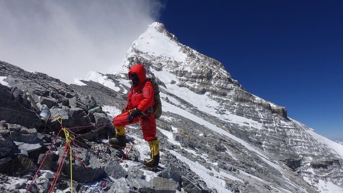 Man in red suit standing on a mountain with the summit of Everest behind him.