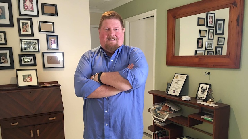 Gary Fitzgerald stands with his arms folded looking at the camera in a room of his home.