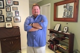 Gary Fitzgerald stands with his arms folded looking at the camera in a room of his home.