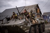 Three Ukrainian soldiers in military uniform sit on top of an armoured military vehicle, moving through a street