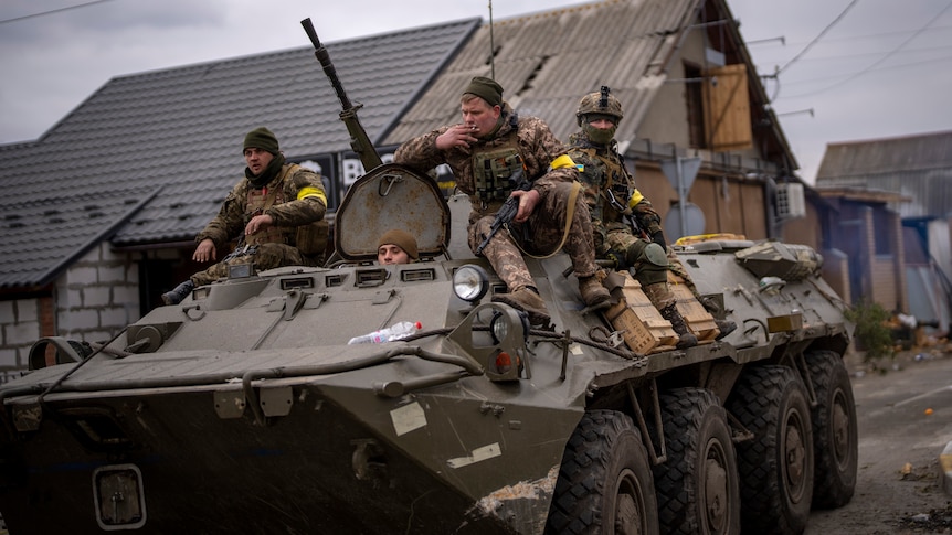 Three Ukrainian soldiers in military uniform sit on top of an armoured military vehicle, moving through a street