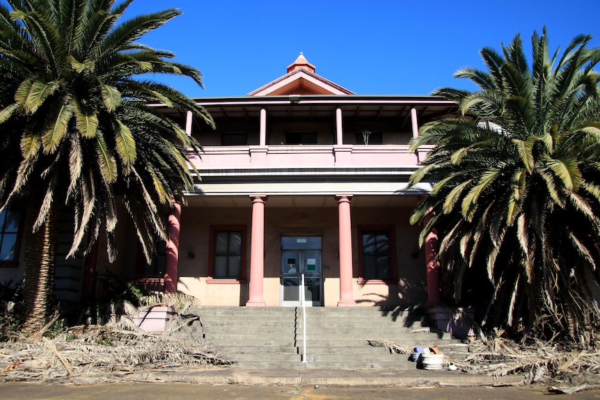 A two storey building with two large palm trees either side and stairs leading up to the building.