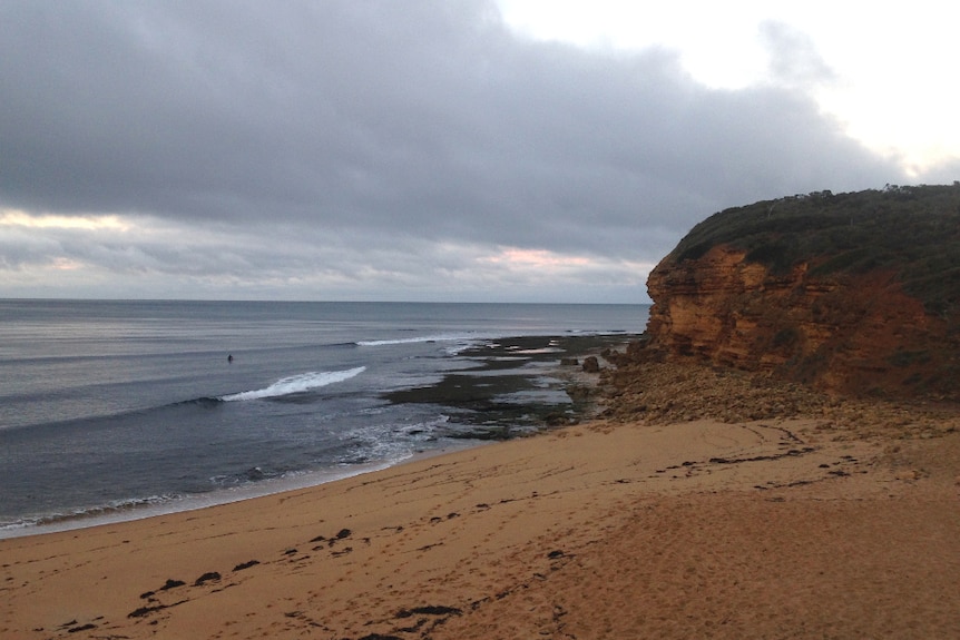 A grey morning at a beach. A single surfer sits out in the lineup.