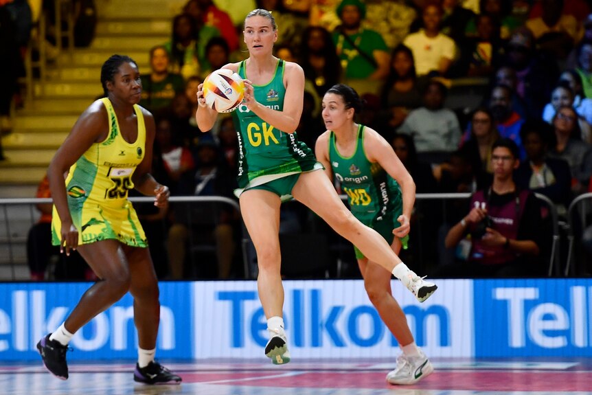 An Australian Diamonds goal keeper catches the ball while off the ground, as a Jamaican goal shooter looks dejected behind her.