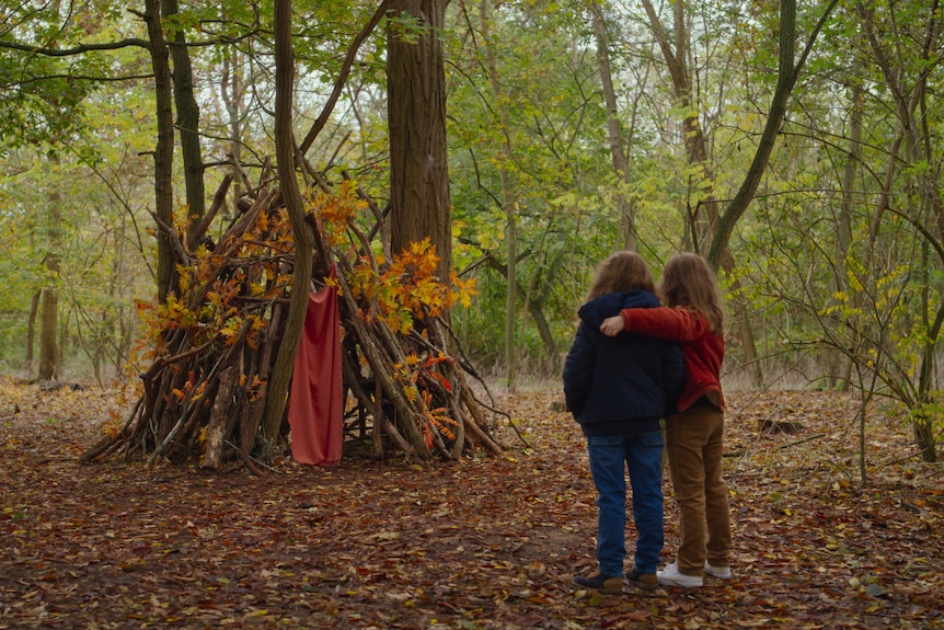 Two young girls huddle together in long-sleeved tops and trousers, looking at a hut made of sticks and leaves in a forest.