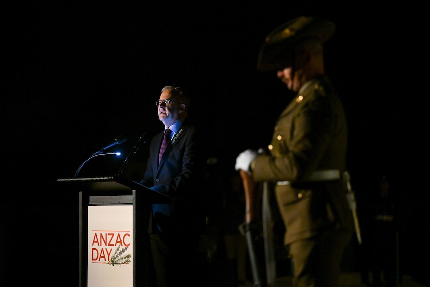 Anthony speaks, surrounded by darkness, a soldier standing nearby under a lamp.