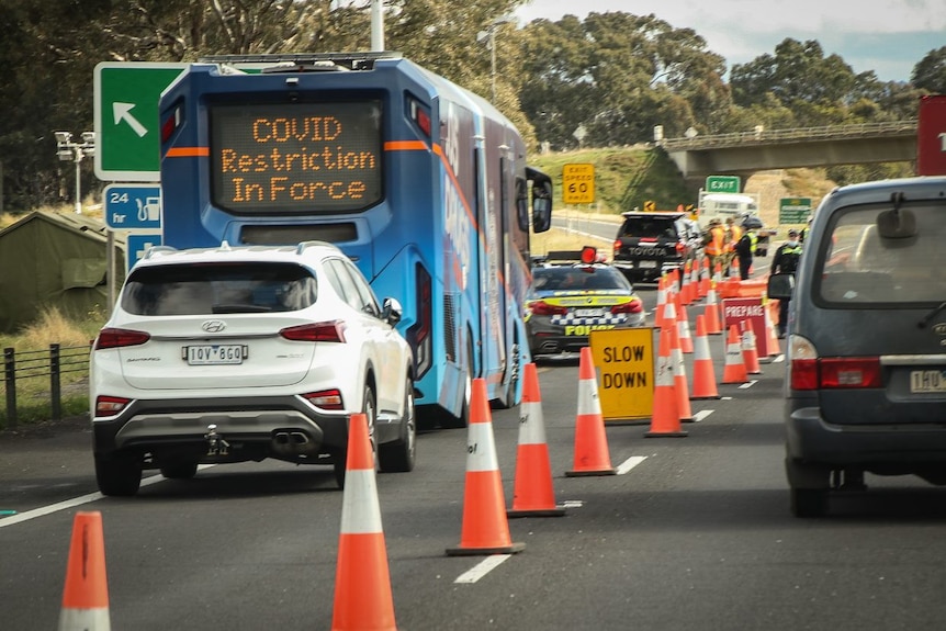 Traffic stopped at roadblock, with a blue bus with a sign saying 'COVID restriction in force'.