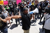 A black man holding a megaphone directs protesters holding placards, "I stand with my black brothers and sisters'.