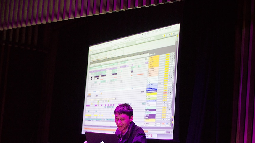 Martin King behind a laptop, on a stage, with a projection of his Ableton project on a screen behind him.