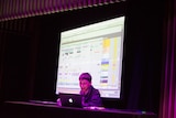 Martin King behind a laptop, on a stage, with a projection of his Ableton project on a screen behind him.