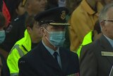A man wearing a suit and medals wears a face mask while others around him don't