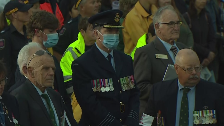 A man wearing a suit and medals wears a face mask while others around him don't