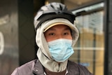 A person wears a rain jacket and helmet over a hood and a face mask on a grey day.