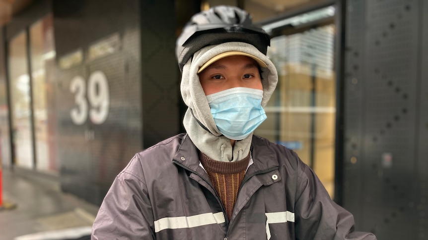 A person wears a rain jacket and helmet over a hood and a face mask on a grey day.