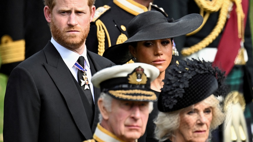 Prince Harry wearing a suit and Meghan wearing a black hat stand behind King Charles and Camilla, Queen Consort