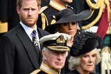 Prince Harry wearing a suit and Meghan wearing a black hat stand behind King Charles and Camilla, Queen Consort