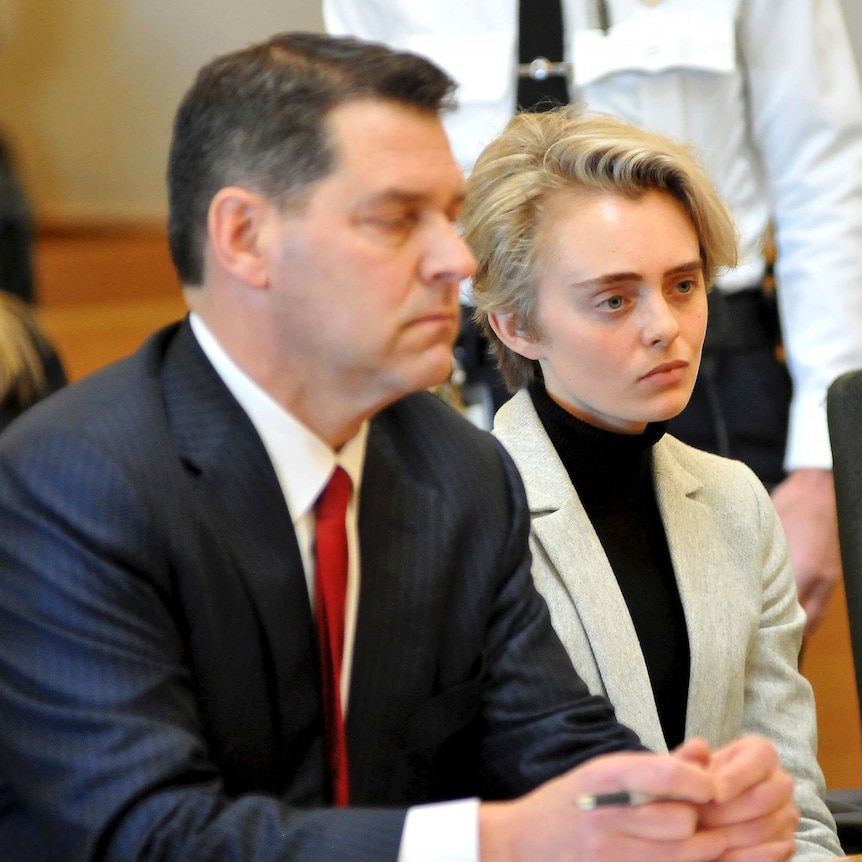 A woman with short blonde hair appears in court sitting next to a man who may be her lawyer.