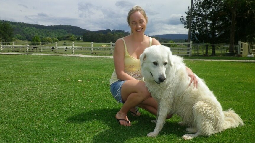 A smiling woman crouches down with her arm laying across a large, white dog, with fields and mountains in the background.