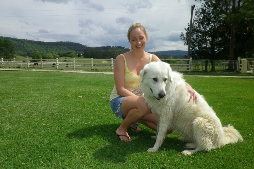 A smiling woman crouches down with her arm laying across a large, white dog, with fields and mountains in the background.