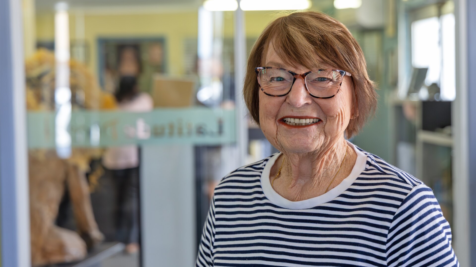 Imagen de una mujer sonriente con gafas. Tiene el pelo corto y está parada frente a un escaparate. 