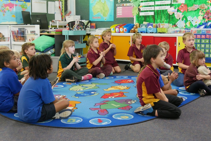 Students in blue, green and maroon uniforms sit on a blue rug in a colourful classroom, clapping and singing.