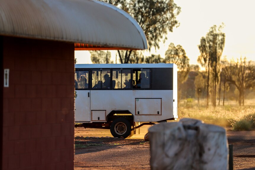 A white dirty bus drives past a toilet block building with sunset and bush in background