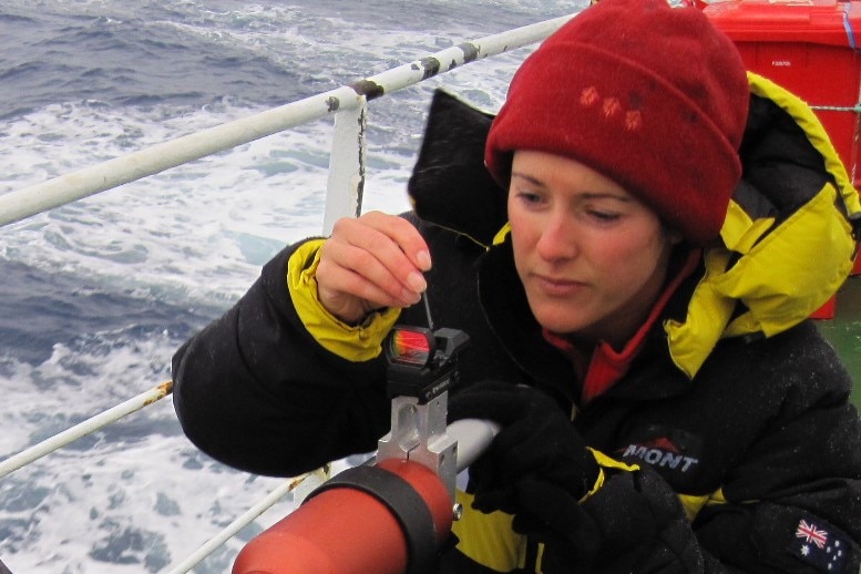 Woman researcher in warm wet weather gear, beanie, crouched at camera instrument aboard a ship at sea 