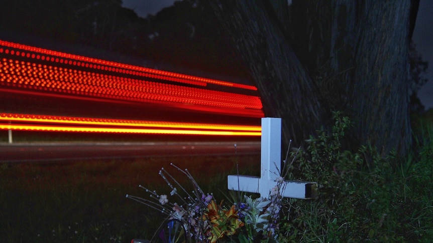 Light streaks at night are seen behind a white cross beside a road.