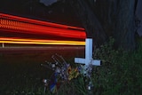 Light streaks at night are seen behind a white cross beside a road.