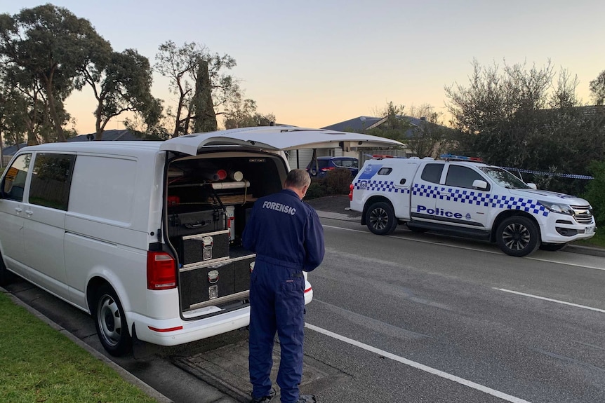 A man in blue overalls with 'FORENSIC' printed on the back retrieves equipment from a white van parked in front of a house.