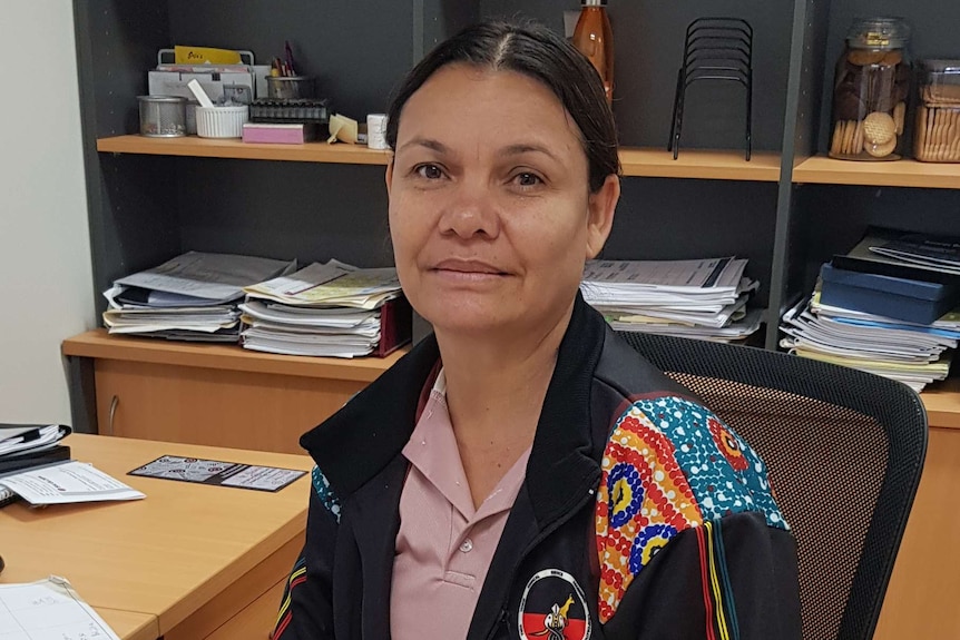 Image of a woman sitting at her desk in an office, wearing a jacket decorated with Indigenous artwork.