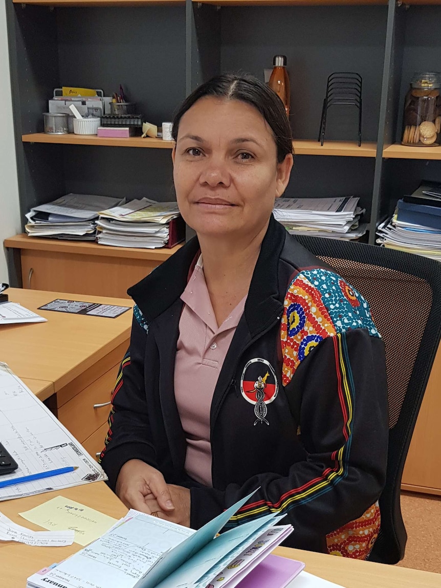 Image of a woman sitting at her desk in an office, wearing a jacket decorated with indigenous artwork.