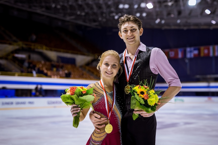 A figure skating pair stand on the ice smiling, holding bouquets and wearing gold medals around their necks after a competition.