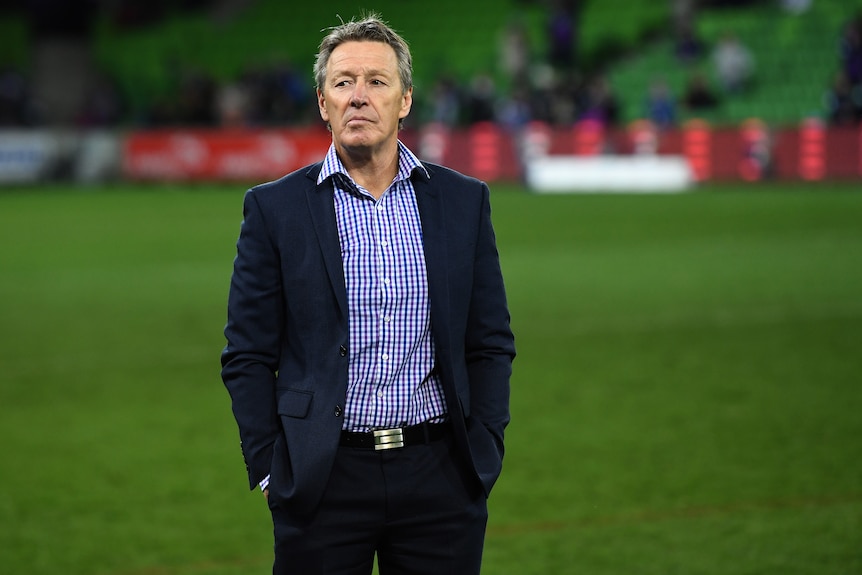 Craig Bellamy stands in a suit in the middle of a stadium with his hands in his pockets