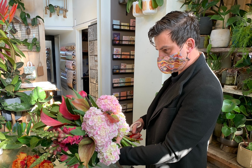 Shane Sipolis, wearing a black jacket and a patterned face mask, holds a bunch of flowers.