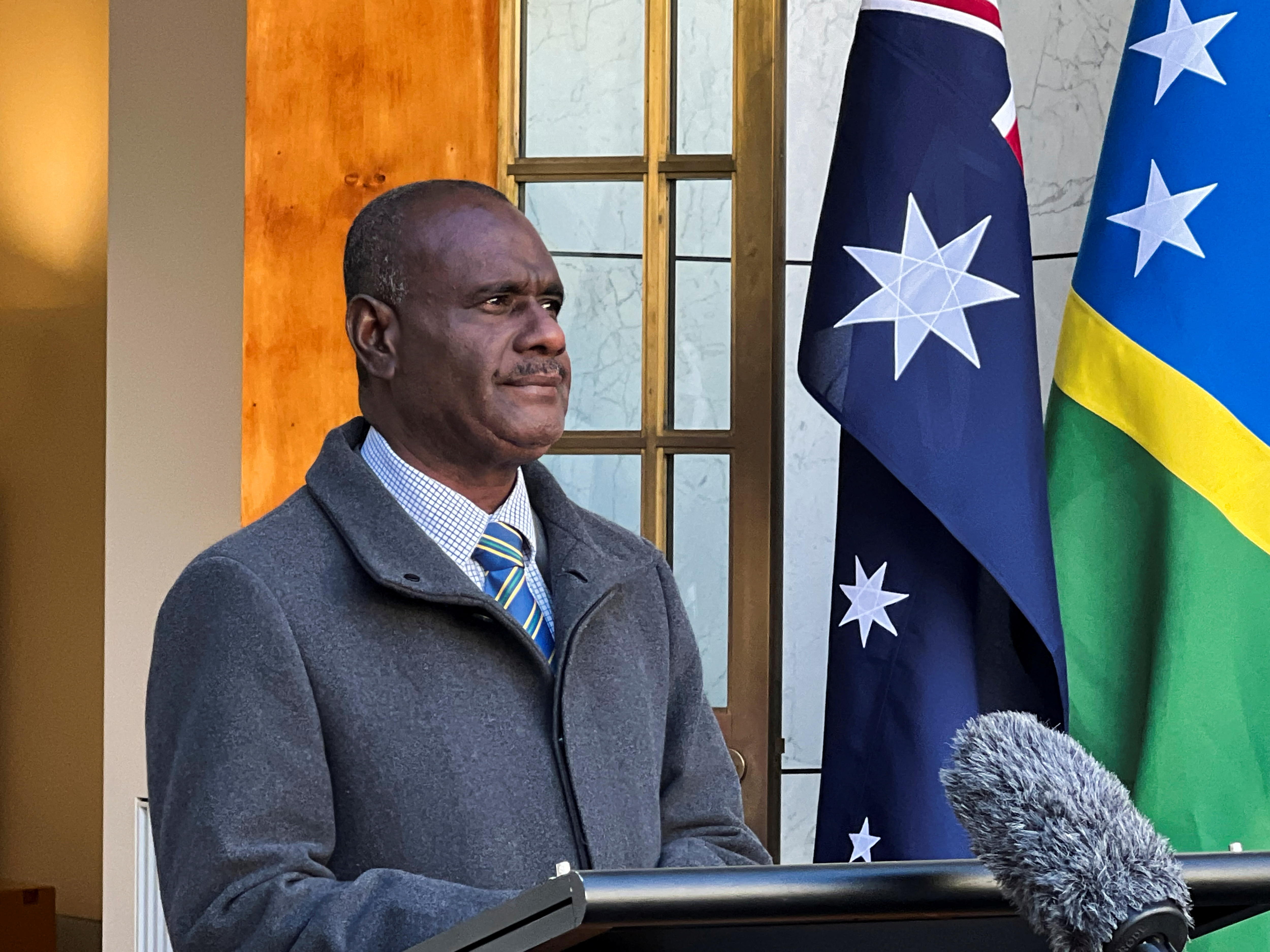 man with very short hair wearing a light blue shirt, striped blue necktie and tailored grey collared jacket stands at mic podium