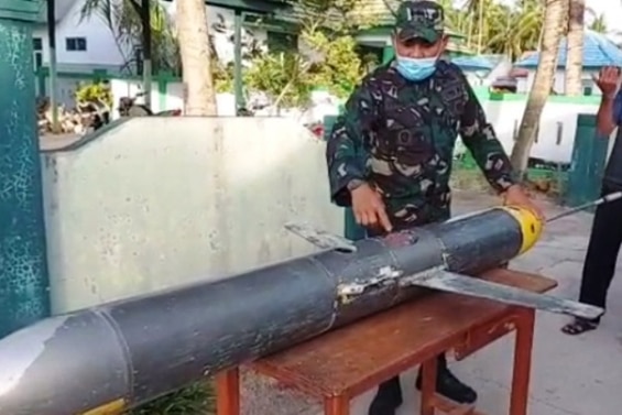 A man in military uniform wearing a facemask stands touching a submarine drone on a table.