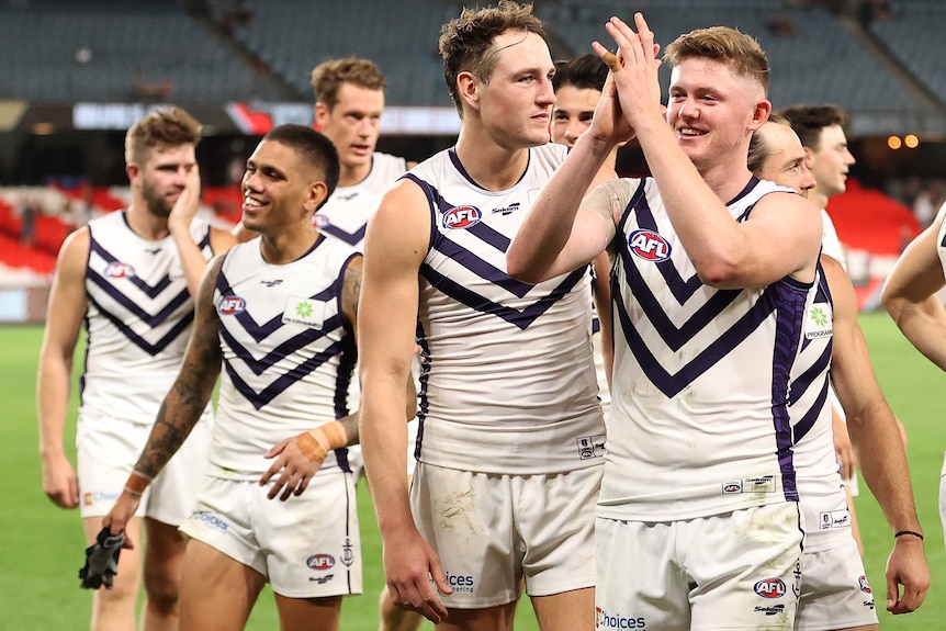A group of smiling AFL players walk off the ground clapping to the crowd after winning a game.