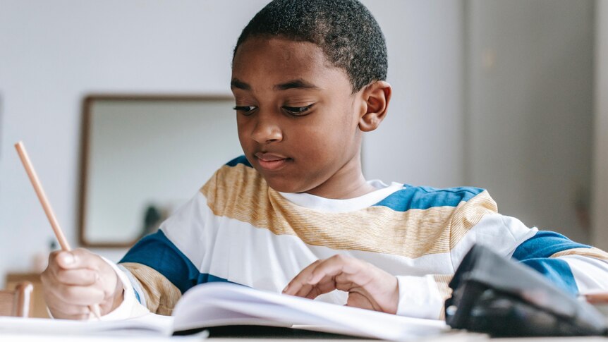 Young boy sits at a table writing something in a workbook, looking at the page.