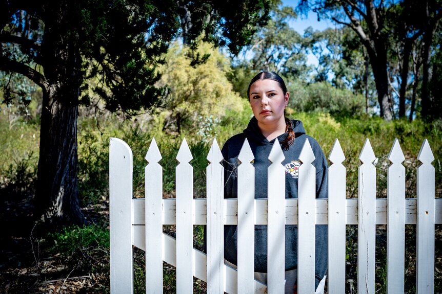Girl wearing a hoodie stands behind white picket fence. 