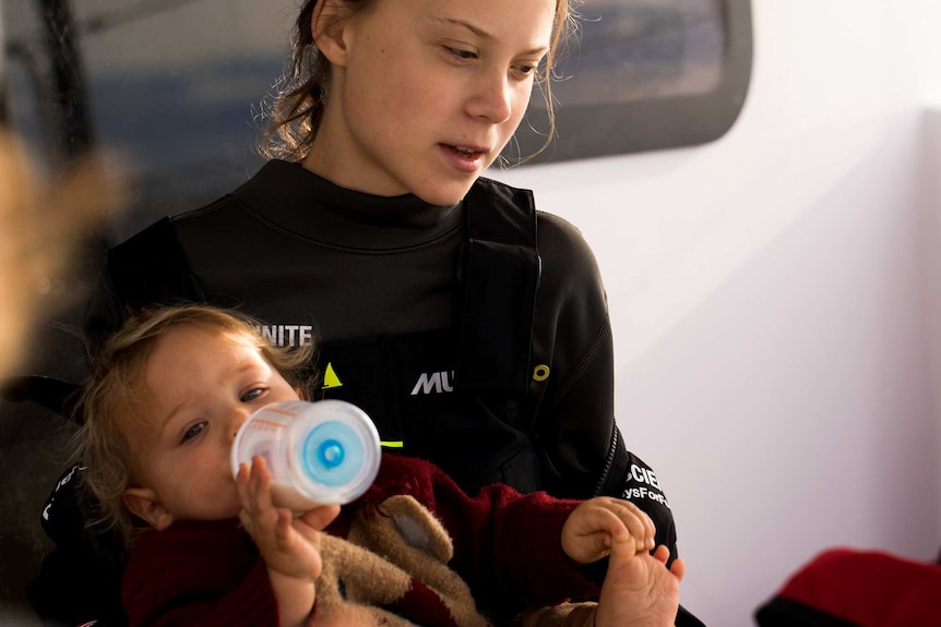 Greta Thunberg wearing a wetsuit and holding a baby as he drinks from a bottle.
